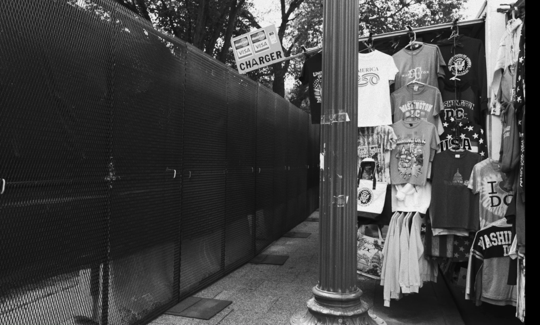 A T-shirt seller idles next to a large black fence